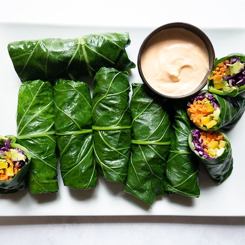 Rectangular white platter topped with veggie-stuffed collard green rolls, served with a small bowl of dipping sauce